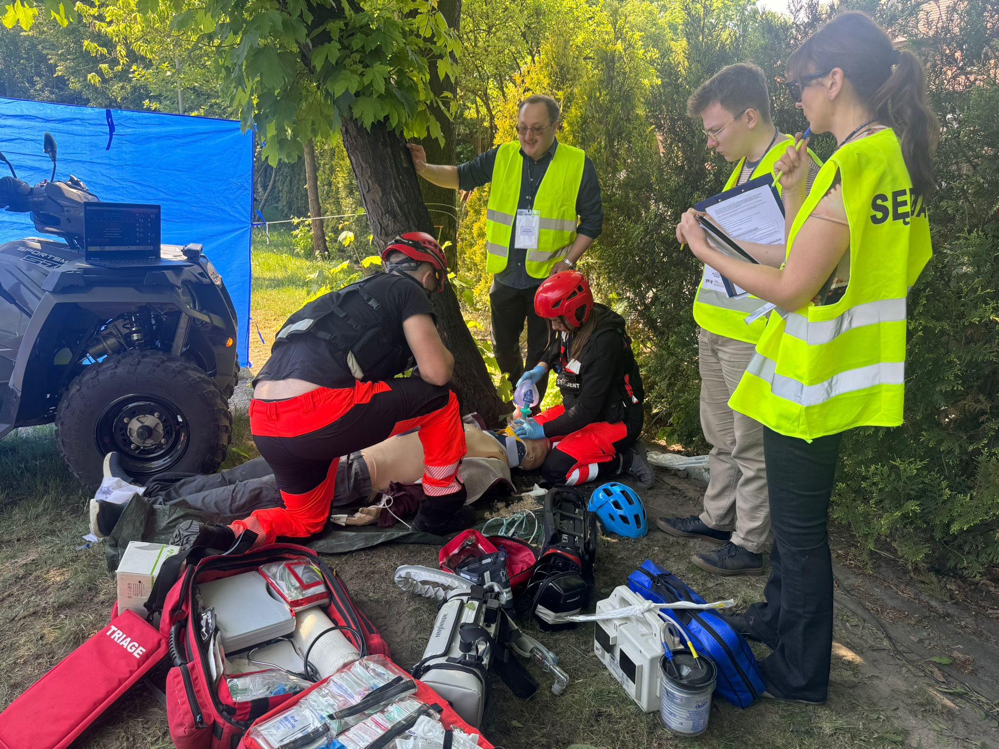 Zespół ratowniczy przeprowadza symulację resuscytacji na manekinie przy quadzie medycznym, podczas gdy sędziowie w kamizelkach obserwują i notują przebieg akcji.