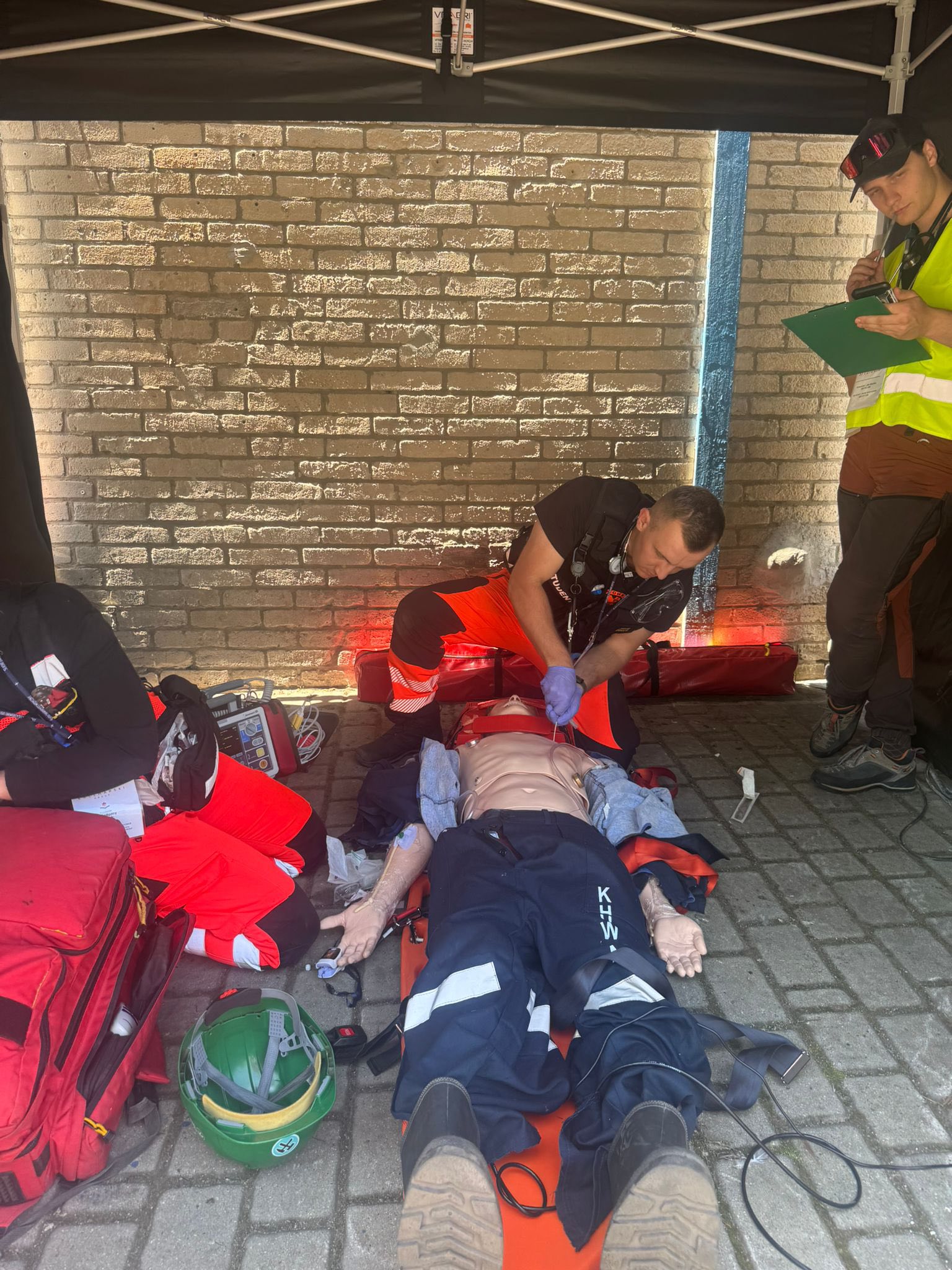 Zespół ratowniczy ćwiczy procedurę zaawansowanych czynności ratunkowych przy manekinie umieszczonym na desce ortopedycznej, w cieniu pod namiotem ratunkowym.