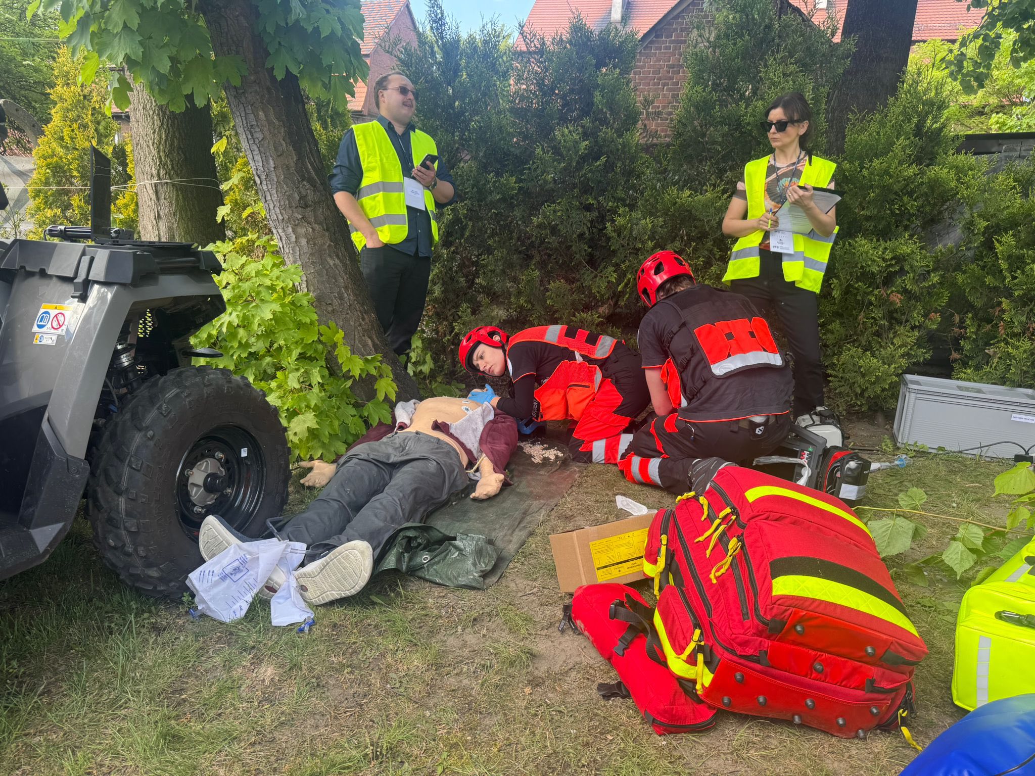 Zespół ratowniczy przeprowadza symulację resuscytacji na manekinie przy quadzie medycznym, podczas gdy sędziowie w kamizelkach obserwują i notują przebieg akcji.