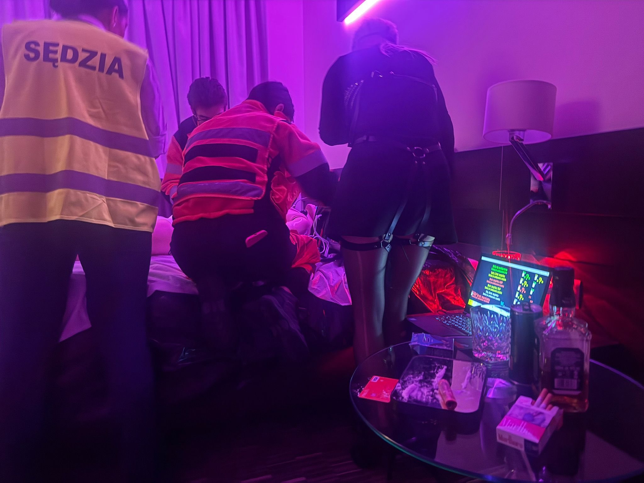 Symulacja interwencji ratowniczej w ciemnym pokoju z oświetleniem UV – ratownicy udzielają pomocy w warunkach imprezy, na stole widoczne rekwizyty imitujące używki, laptop z symulacją parametrów pacjenta.