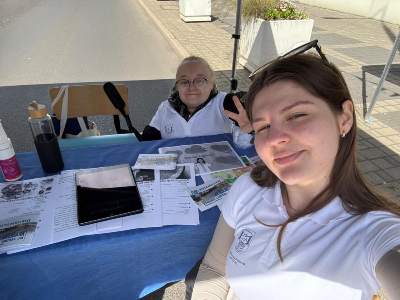 Dwie młode kobiety w białych koszulkach z logo uczelni uśmiechają się do selfie przy stole z materiałami informacyjnymi i planszami edukacyjnymi. W tle namiot, pod którym znajduje się stoisko informacyjne.