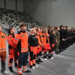 Ustawiona w szeregu grupa uczestników w hali sportowo-widowiskowej podczas oficjalnej części wydarzenia. Po lewej stronie stoją studenci ratownictwa medycznego w pomarańczowo-czarnych strojach z elementami odblaskowymi. Obok nich widoczni są uczniowie lub członkowie służb mundurowych w czarnych i zielonych uniformach. W tle widać puste trybuny oraz kilku funkcjonariuszy obserwujących uroczystość. Zdjęcie przedstawia moment zbiórki lub rozpoczęcia zawodów lub ćwiczeń służb ratunkowych i mundurowych.