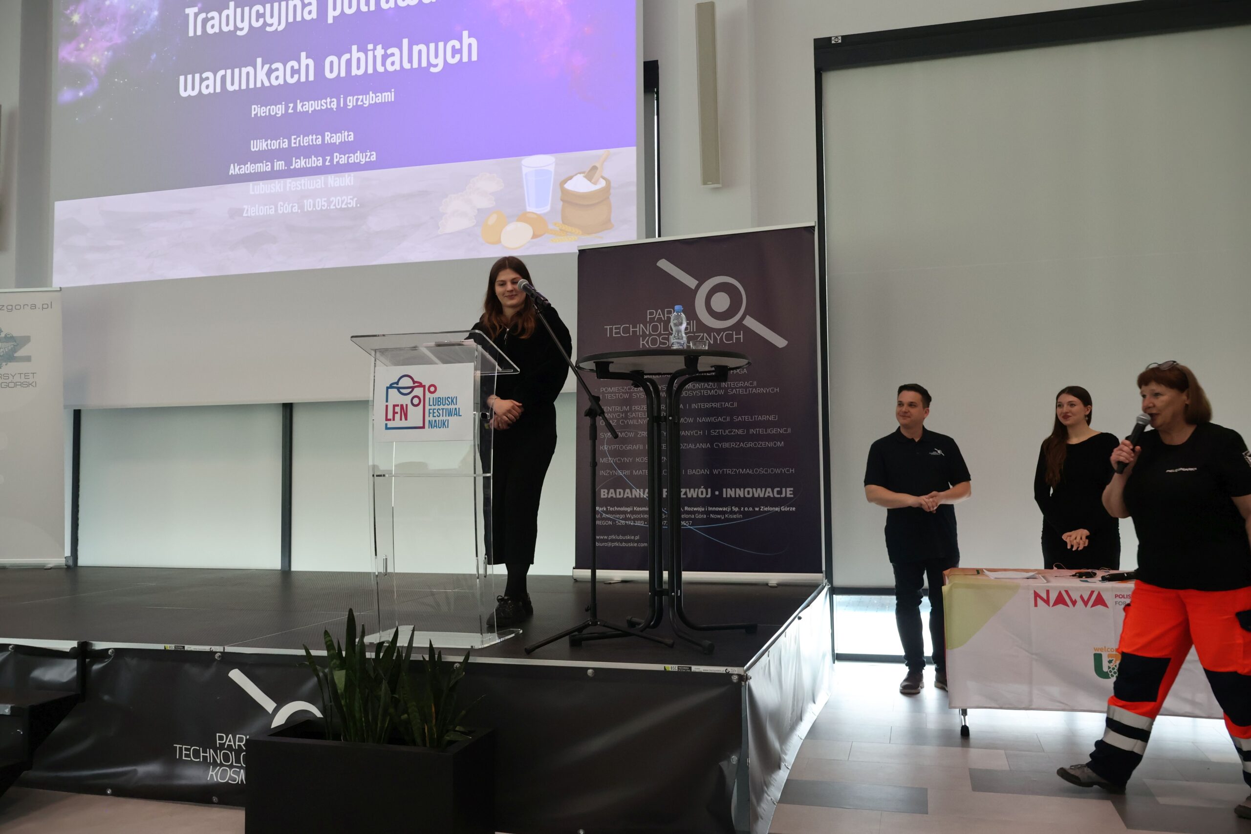 Studentka Wiktoria Rapita z Wydziału Nauk o Zdrowiu prezentuje referat pt. „Tradycyjna potrawa w warunkach orbitalnych – pierogi z kapustą i grzybami” podczas Lubuskiego Festiwalu Nauki. Na scenie podium z logo LFN, w tle slajd z tytułem prelekcji i grafiką kosmiczną.