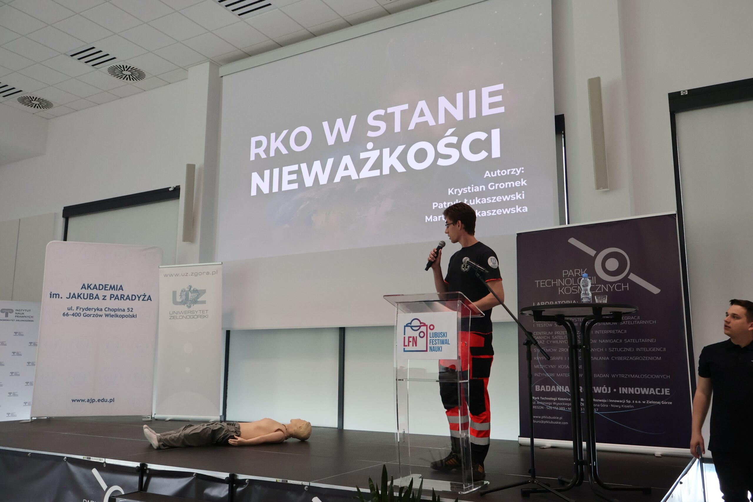 Student Wydziału Nauk o Zdrowiu prezentuje temat „RKO w stanie nieważkości” na scenie Lubuskiego Festiwalu Nauki. W tle plansza z tytułem prezentacji oraz logotypy uczelni. Obok prelegenta znajduje się manekin do ćwiczeń resuscytacyjnych.