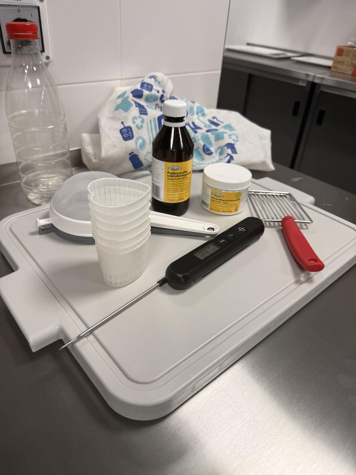 Zestaw przyborów do produkcji sera na blacie kuchennym – termometr kuchenny, sitka, pojemniczki serowarskie, podpuszczka mikrobiologiczna i akcesoria.