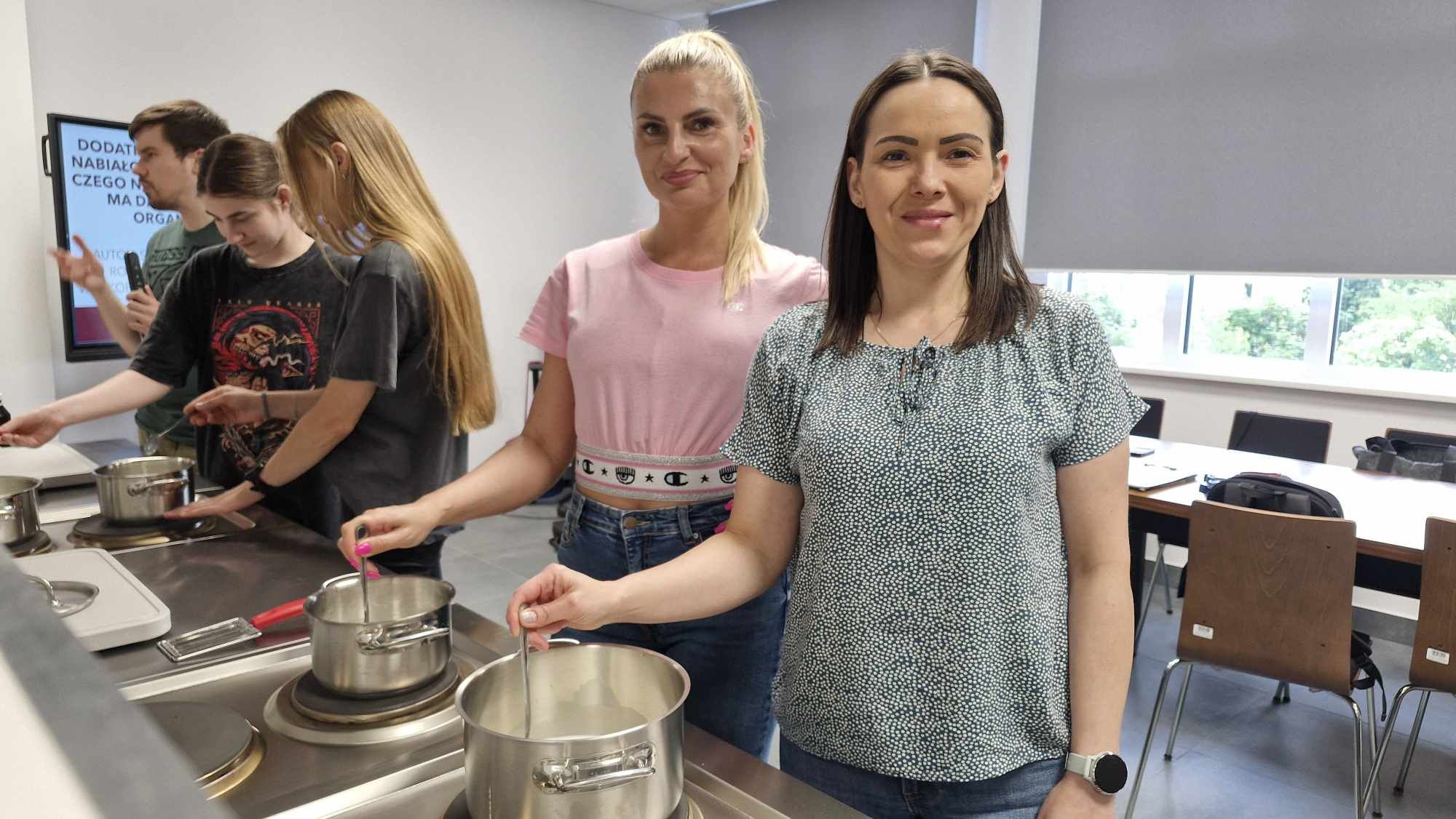 Studenci II roku dietetyki mieszają mleko w garnkach na zajęciach praktycznych – uśmiechnięte studentki stojące przy stanowisku kuchennym.