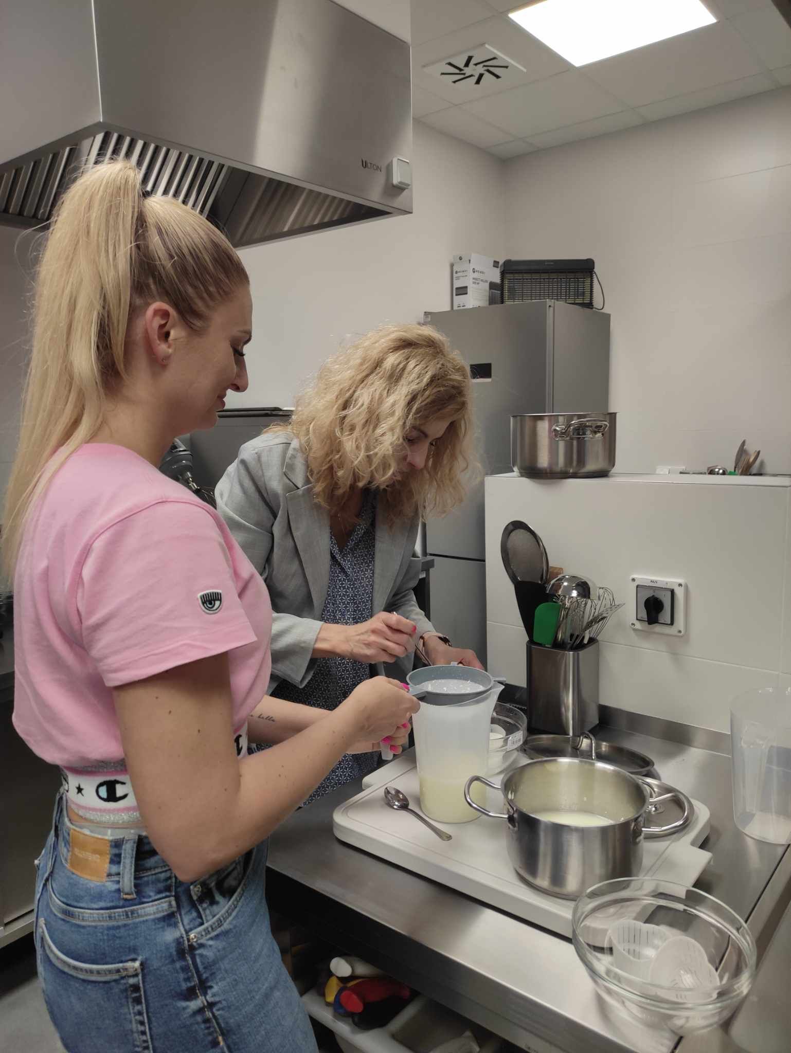 Studentki filtrują serwatkę do wysokiego naczynia – przygotowanie do formowania serków.