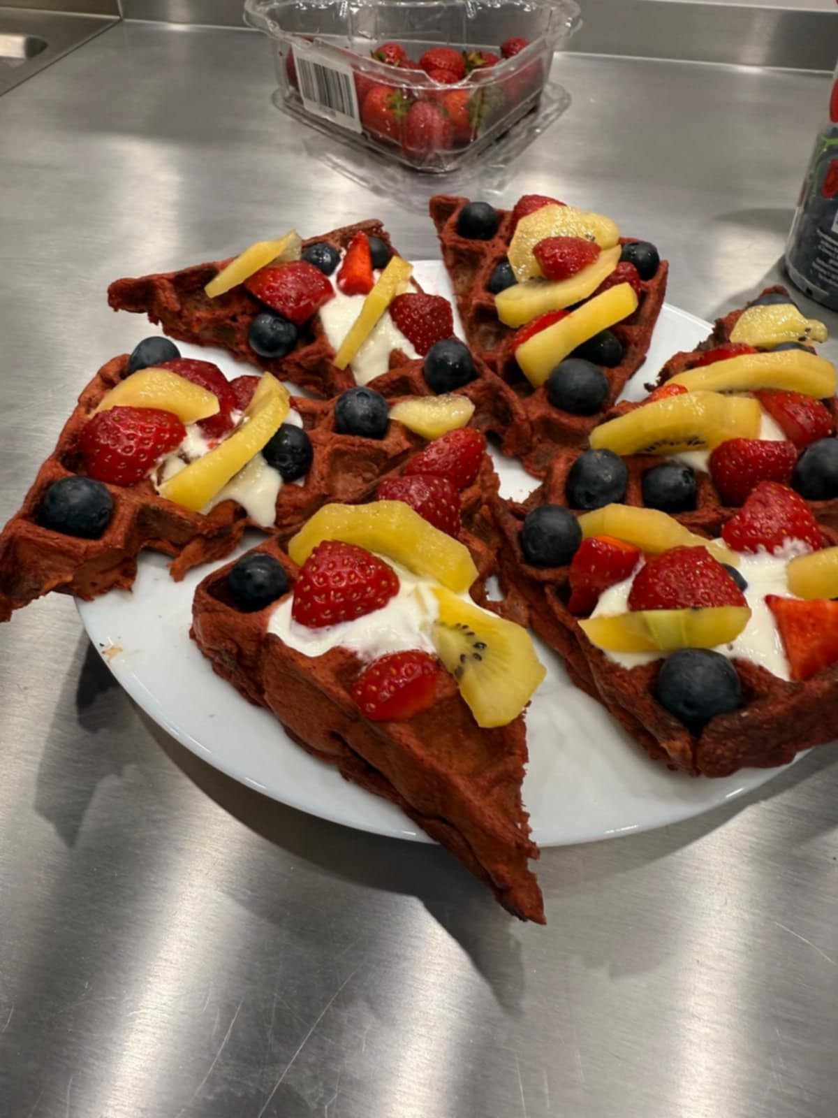 Talerz z kawałkami czerwonych gofrów podanych ze śmietaną, plasterkami kiwi, truskawkami, borówkami i brzoskwinią. Gofry ułożone są estetycznie, tworząc apetyczne i kolorowe danie.
