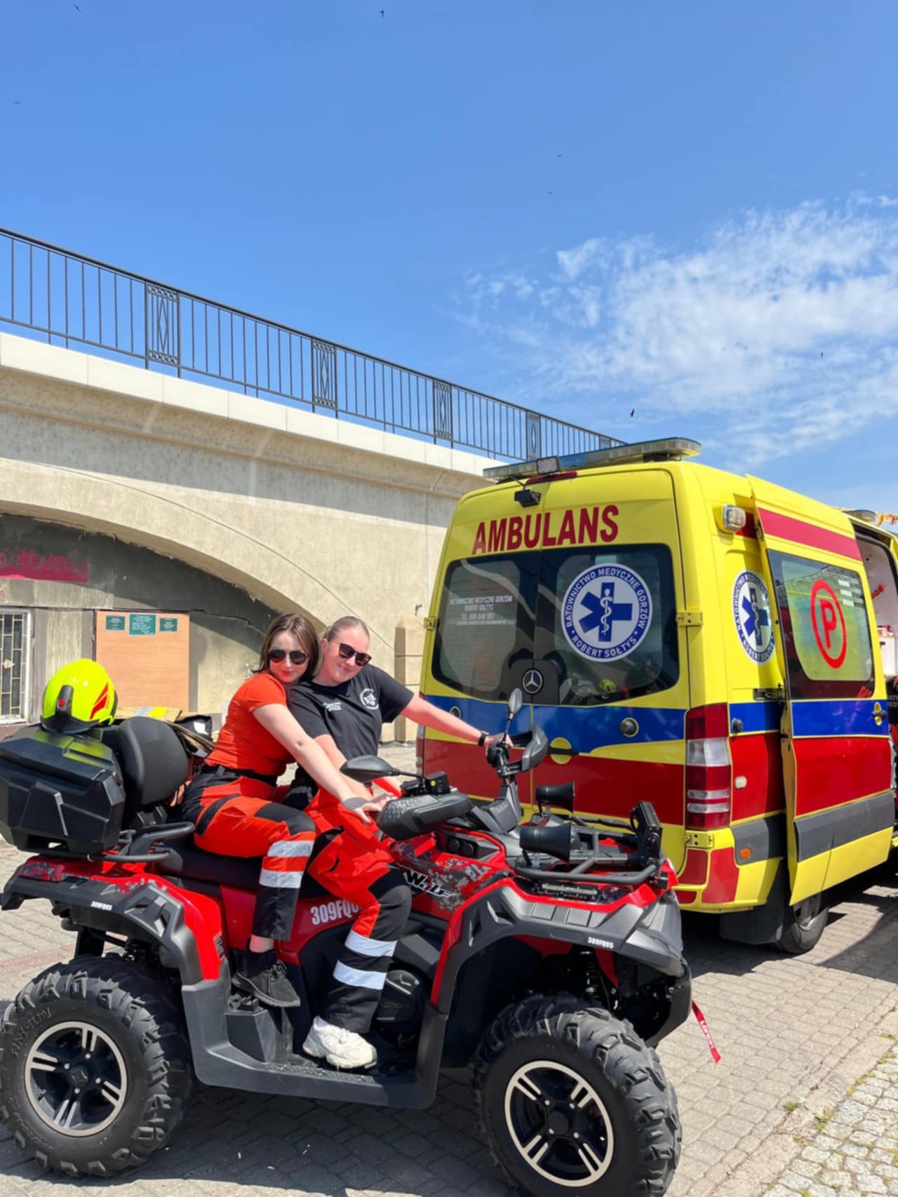 Dwie osoby w odzieży ratowniczej siedzą na dużym, czerwonym quadzie ratowniczym, zaparkowanym obok żółto-czerwonego ambulansu. Uśmiechają się do kamery, a w tle widoczny jest mur wiaduktu i błękitne niebo z delikatnymi chmurami. Na quadzie umieszczony jest kask ratowniczy oraz oznaczenia służb ratowniczych. Zdjęcie wykonane podczas plenerowego wydarzenia.