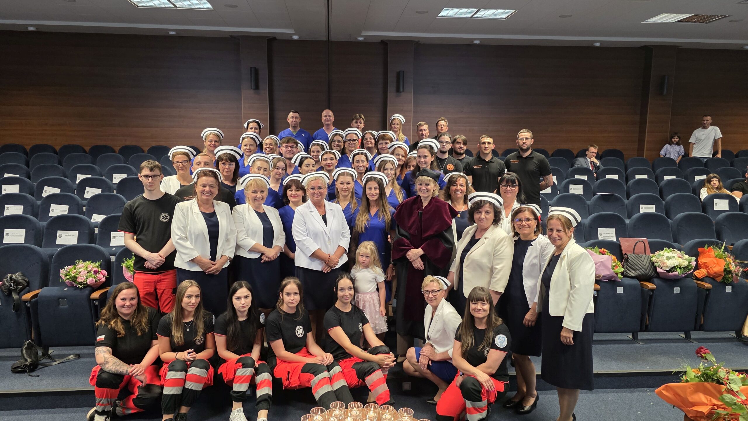 Uroczyste zdjęcie grupowe wykonane w auli uczelni, przedstawiające absolwentów kierunku pielęgniarstwo w czepkach i granatowych strojach oraz absolwentów ratownictwa medycznego w czerwono-czarnych spodniach i czarnych koszulkach, wykładowczynie w eleganckich strojach oraz dziekana w todze akademickiej. Zdjęcie ukazuje wspólne świętowanie zakończenia roku akademickiego.