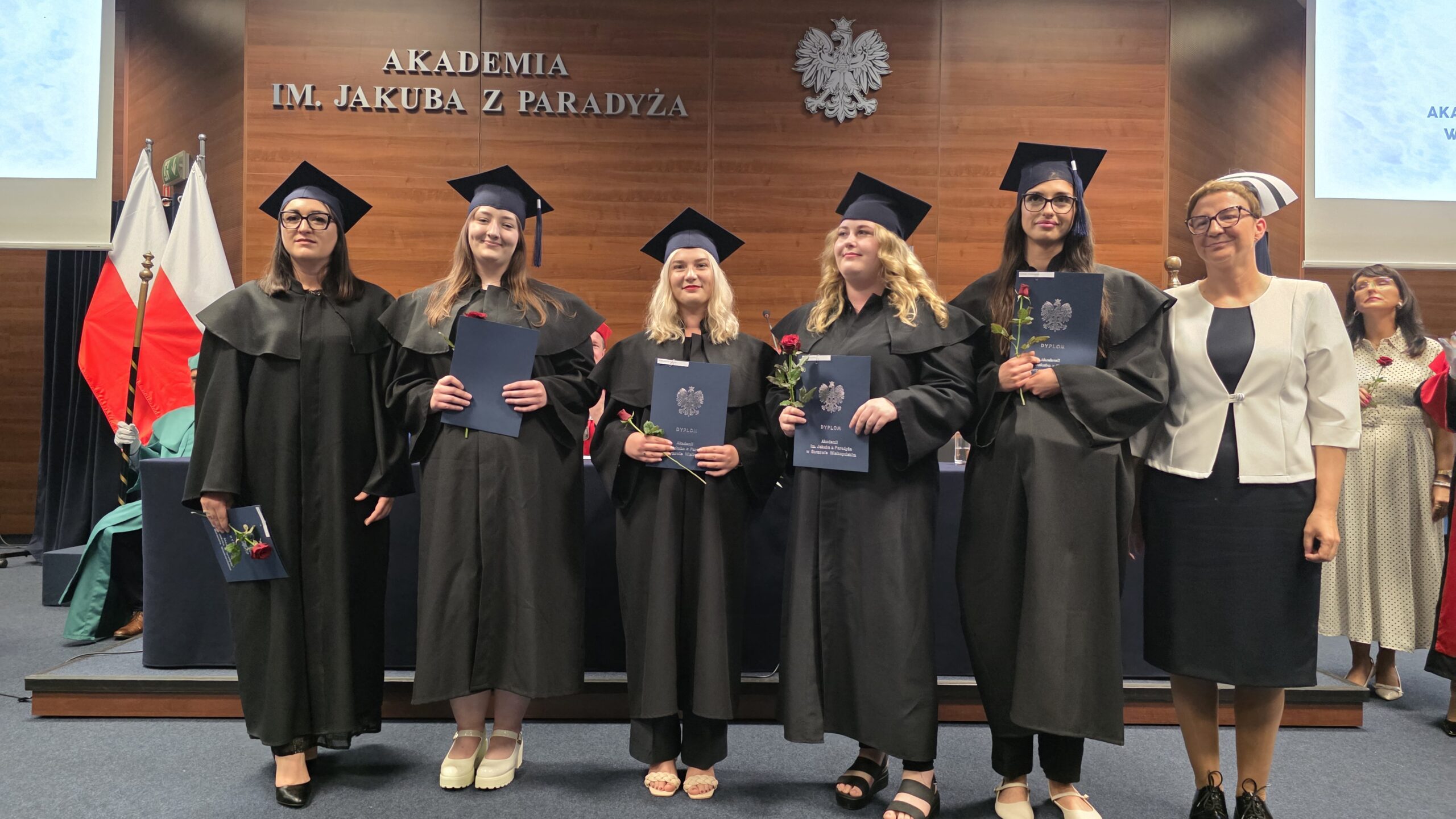 Pięć absolwentek Akademii im. Jakuba z Paradyża ubranych w togi i birety pozuje z dyplomami i czerwonymi różami, stojąc na tle drewnianej ściany z godłem Polski i napisem „AKADEMIA IM. JAKUBA Z PARADYŻA”. Obok nich stoi wykładowczyni w białym żakiecie i czepku pielęgniarskim. W tle widoczne są flagi Polski oraz osoby w togach i strojach akademickich. Zdjęcie wykonano podczas uroczystości wręczenia dyplomów 11 lipca 2025 roku.