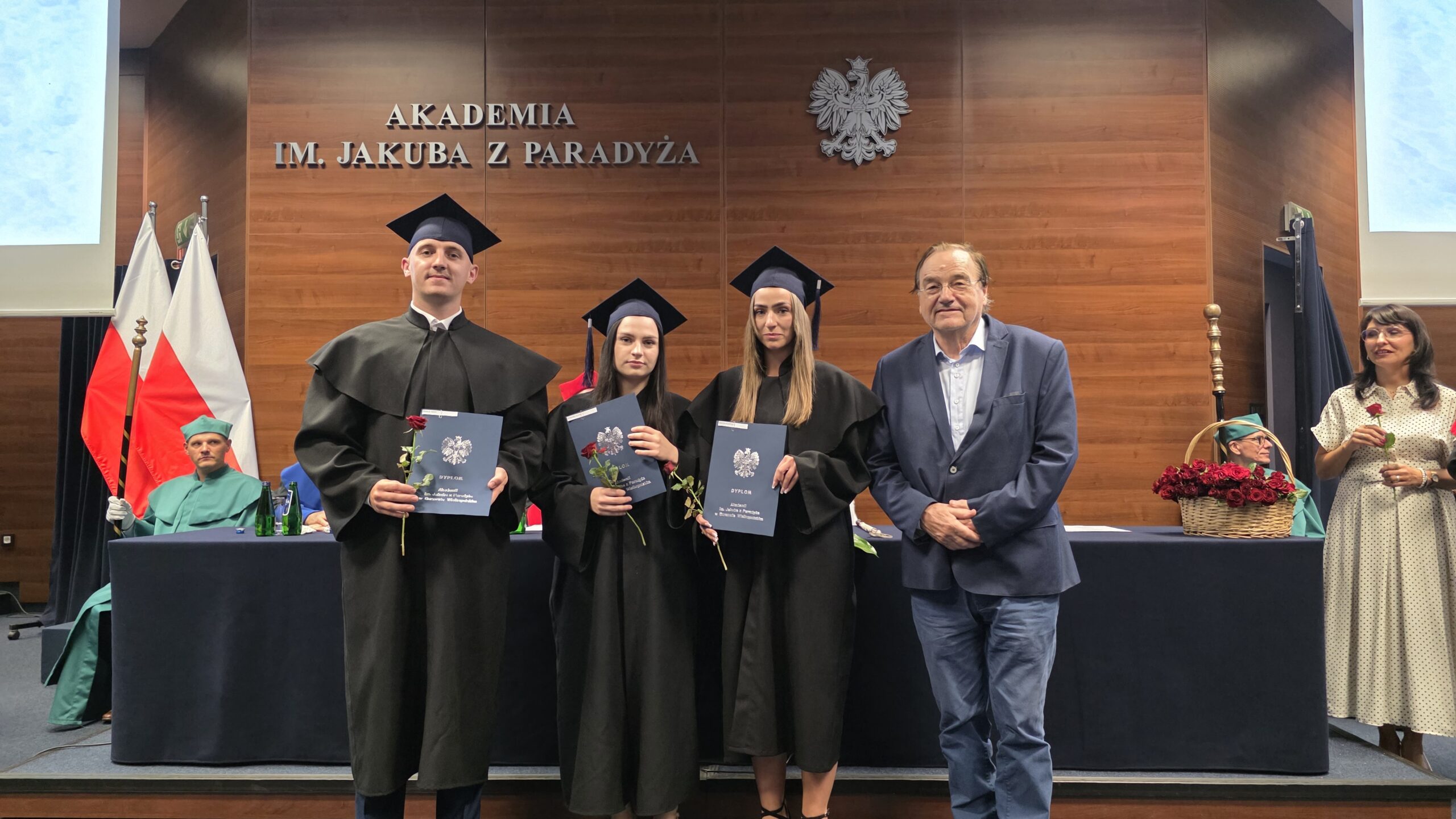 Trzech absolwentów Akademii im. Jakuba z Paradyża – jeden mężczyzna i dwie kobiety – pozuje na scenie w togach i biretach, trzymając dyplomy oraz czerwone róże. Obok nich stoi profesor w niebieskiej marynarce. W tle widoczne są flagi Polski, godło, osoby w strojach akademickich oraz kosz wypełniony różami. Zdjęcie wykonano podczas uroczystości wręczenia dyplomów.