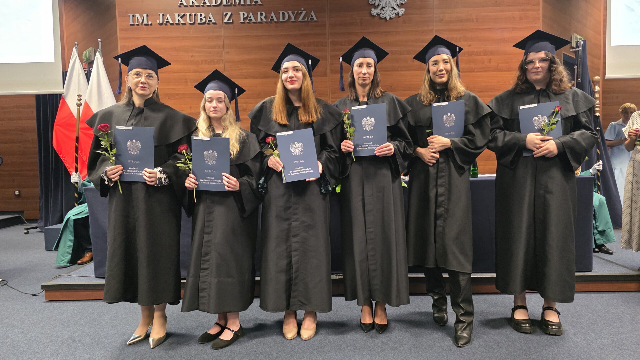 Sześć absolwentek Akademii im. Jakuba z Paradyża pozuje na scenie w czarnych togach i biretach, trzymając w dłoniach dyplomy oraz czerwone róże. W tle widoczne są flagi Polski, godło oraz drewniana ściana z napisem „AKADEMIA IM. JAKUBA Z PARADYŻA”. Zdjęcie wykonano podczas oficjalnej uroczystości wręczenia dyplomów.