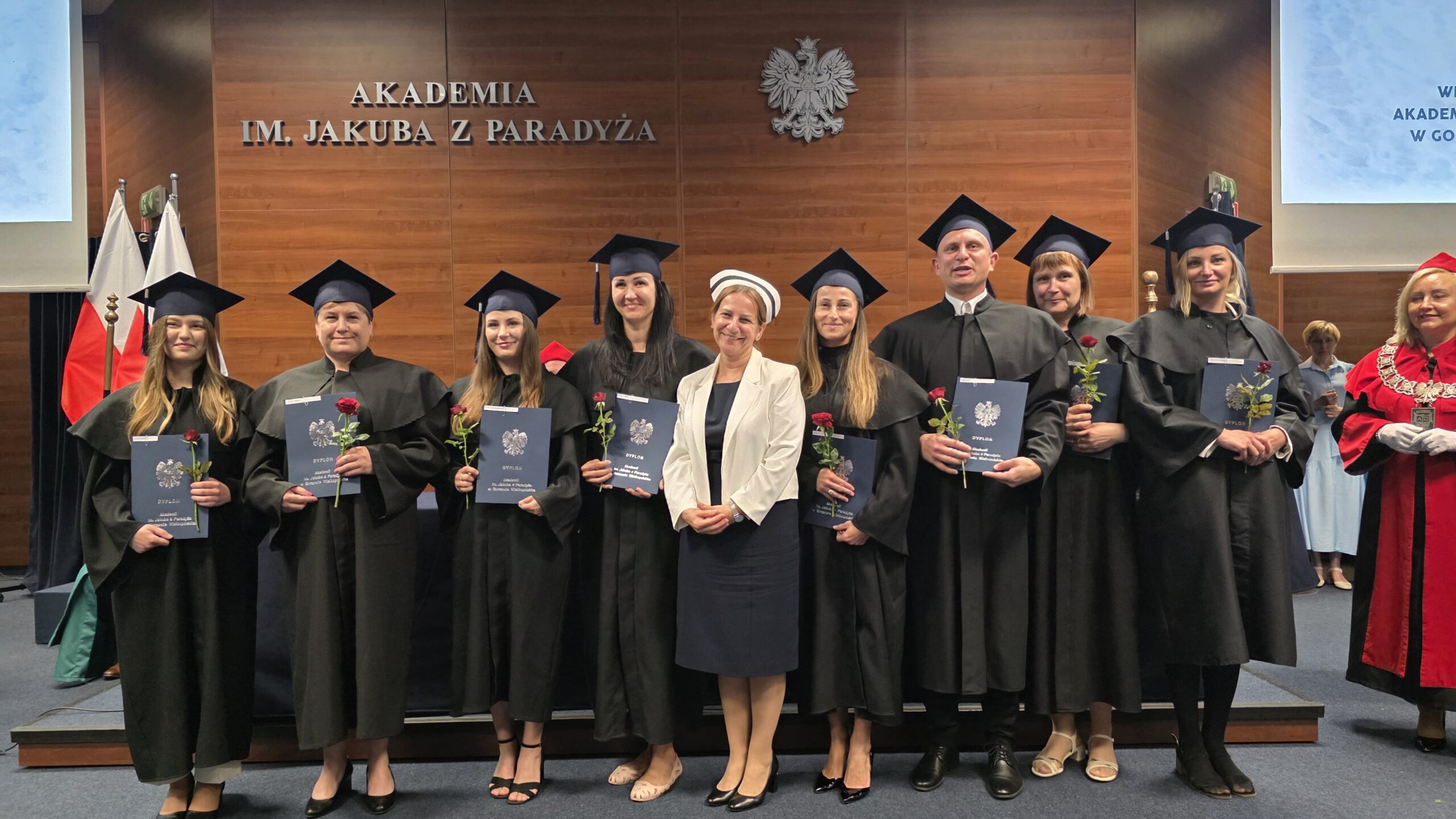 Ośmioro absolwentów Akademii im. Jakuba z Paradyża w togach i biretach pozuje z dyplomami i czerwonymi różami na tle drewnianej ściany z napisem „AKADEMIA IM. JAKUBA Z PARADYŻA” oraz godłem Polski. W centrum stoi wykładowczyni w białym żakiecie i pielęgniarskim czepku. Po prawej stronie widoczna jest Prorektor uczelni w czerwonej todze z insygniami. Zdjęcie wykonane podczas uroczystego wręczenia dyplomów.