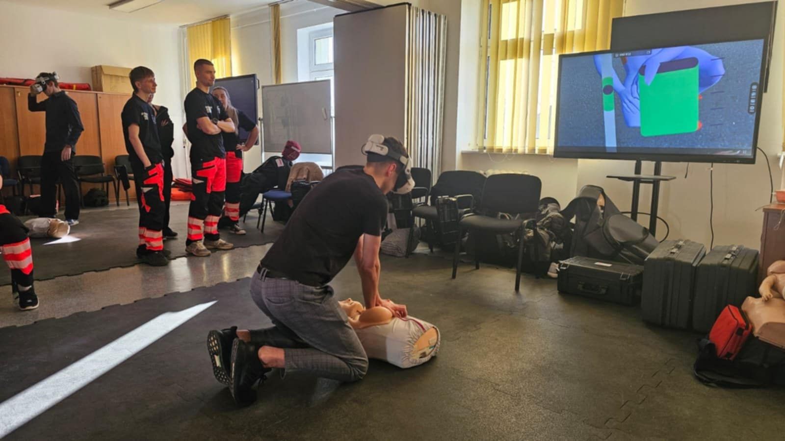 „Student w goglach VR wykonuje uciśnięcia klatki piersiowej na fantomie do resuscytacji, podczas gdy pozostali uczestnicy warsztatów obserwują ćwiczenia w sali dydaktycznej.”