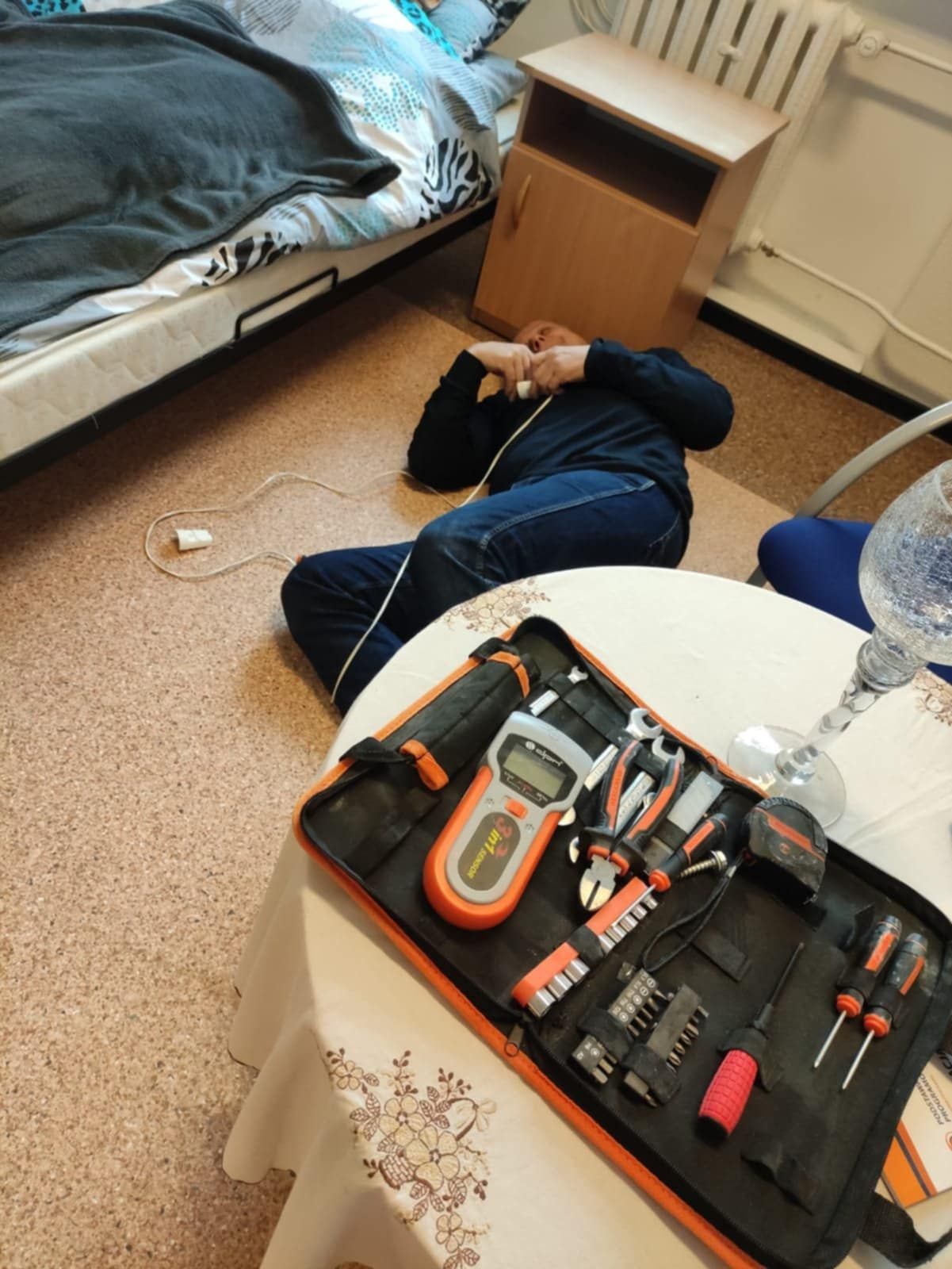Osoba leży na podłodze w pokoju obok łóżka, trzymając w ręku przewód elektryczny. Na stole na pierwszym planie znajduje się otwarty zestaw narzędzi.