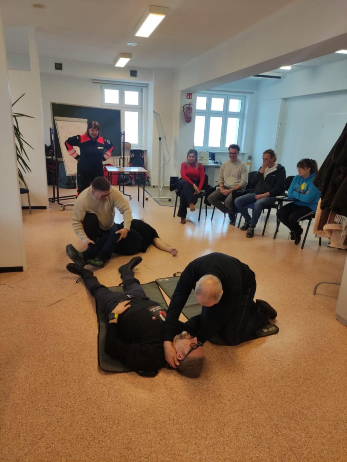 Grupa osób uczestniczy w ćwiczeniach z pierwszej pomocy. Dwóch uczestników wykonuje czynności ratunkowe na osobach pozorujących poszkodowanych, podczas gdy reszta grupy obserwuje instruktaż.