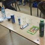 Na stole w klasie ustawiono różne produkty spożywcze zawierające cukier, m.in. napój energetyczny, baton, galaretkę, żelki i słodki jogurt. Obok stoją puste szklane słoiczki oraz pojemnik z cukrem używany do pokazu ilości cukru w produktach.