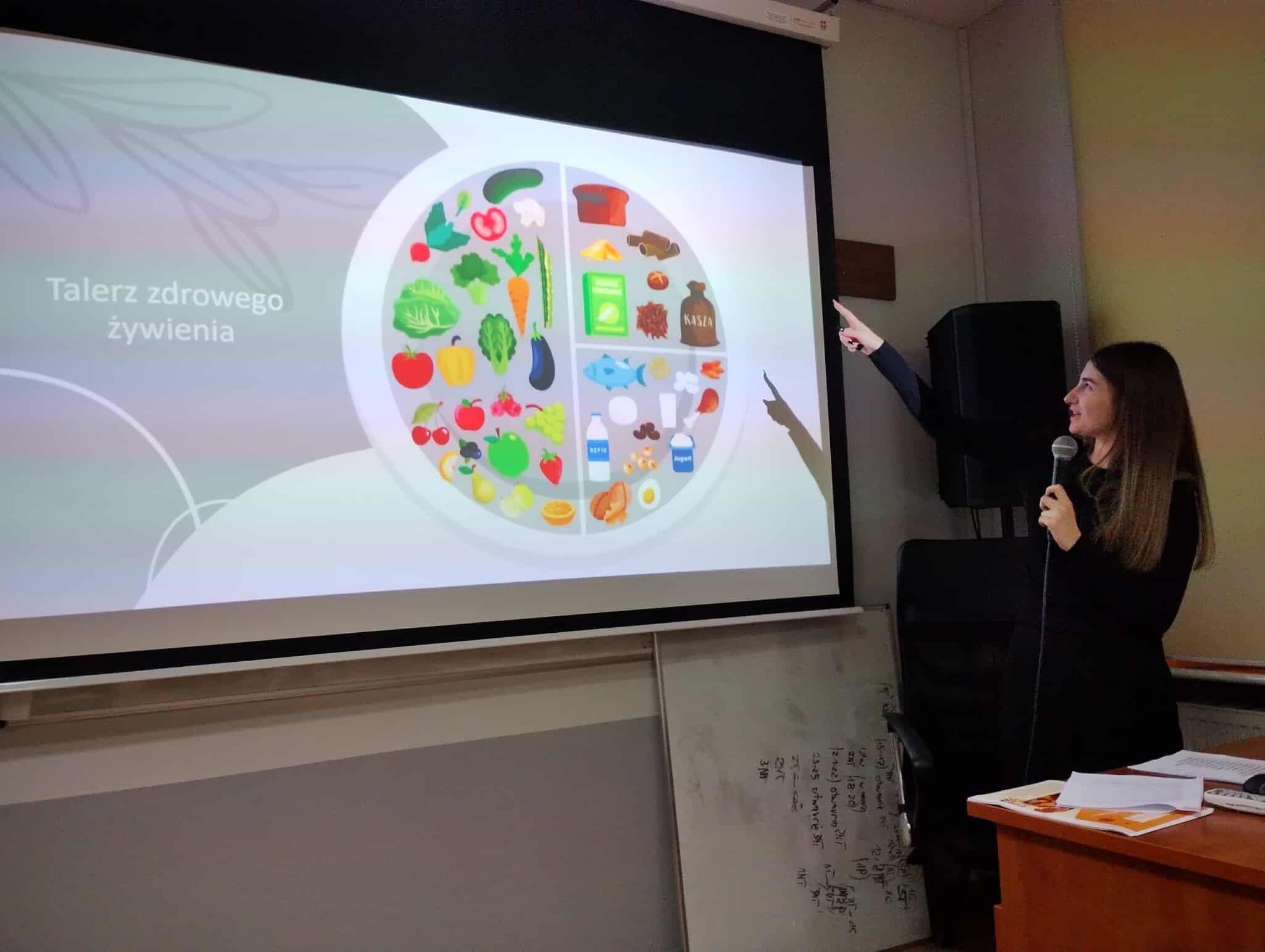 Prowadząca wskazuje na slajd przedstawiający „Talerz zdrowego żywienia”, na którym zobrazowano grupy produktów spożywczych. Stoi obok ekranu, trzymając mikrofon.