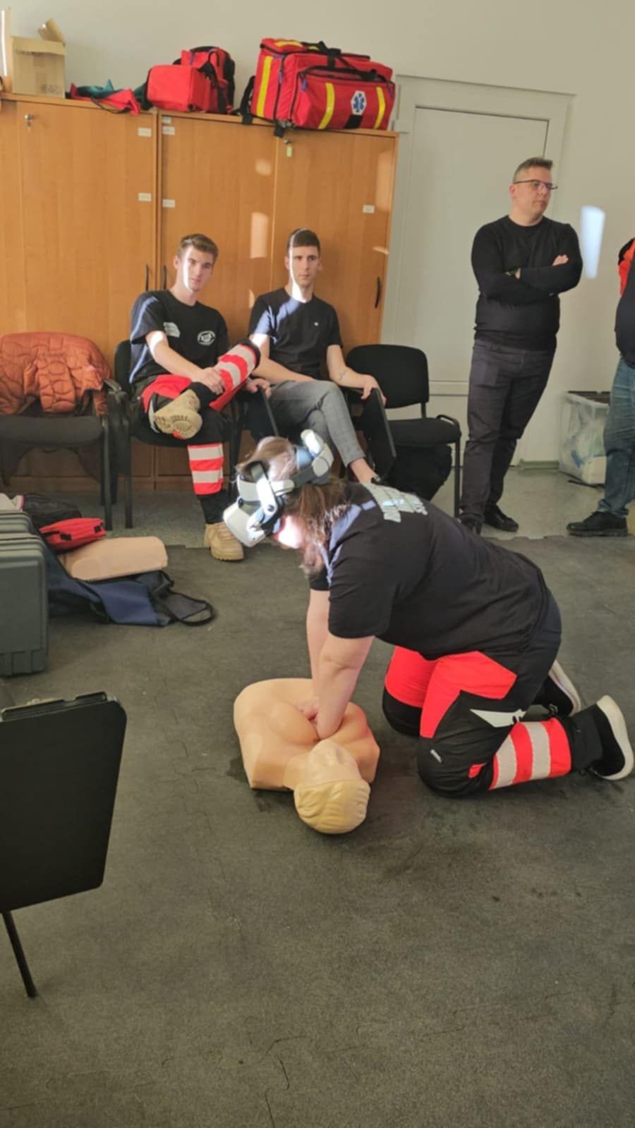 „Uczestniczka warsztatów w goglach VR ćwiczy resuscytację krążeniowo-oddechową na fantomie. W tle kilku studentów siedzi i obserwuje przebieg zajęć.”