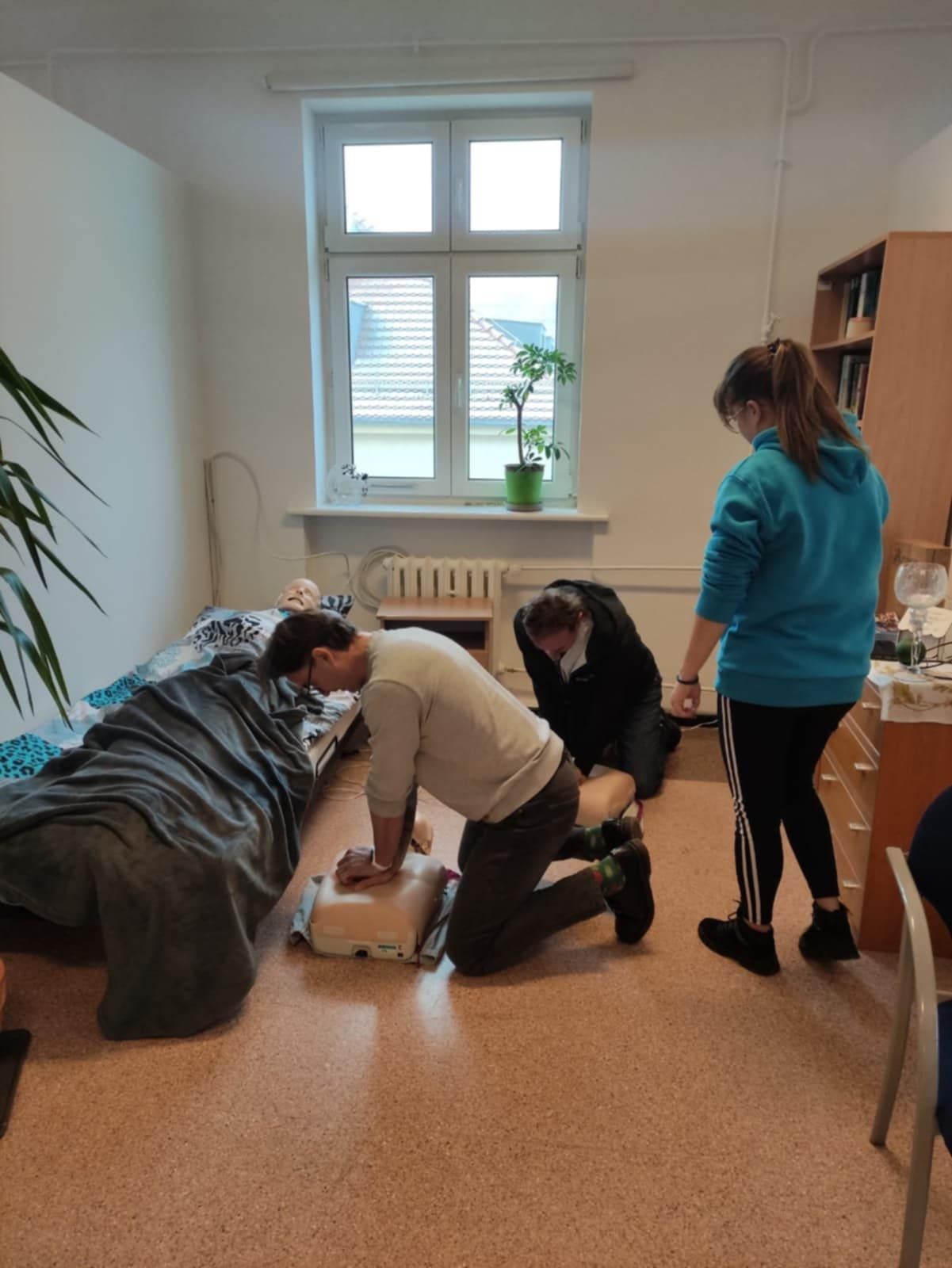 Uczestnicy warsztatów ćwiczą resuscytację krążeniowo-oddechową na fantomie w pokoju symulacyjnym. Jedna osoba wykonuje uciśnięcia klatki piersiowej, a pozostali obserwują przebieg ćwiczenia.