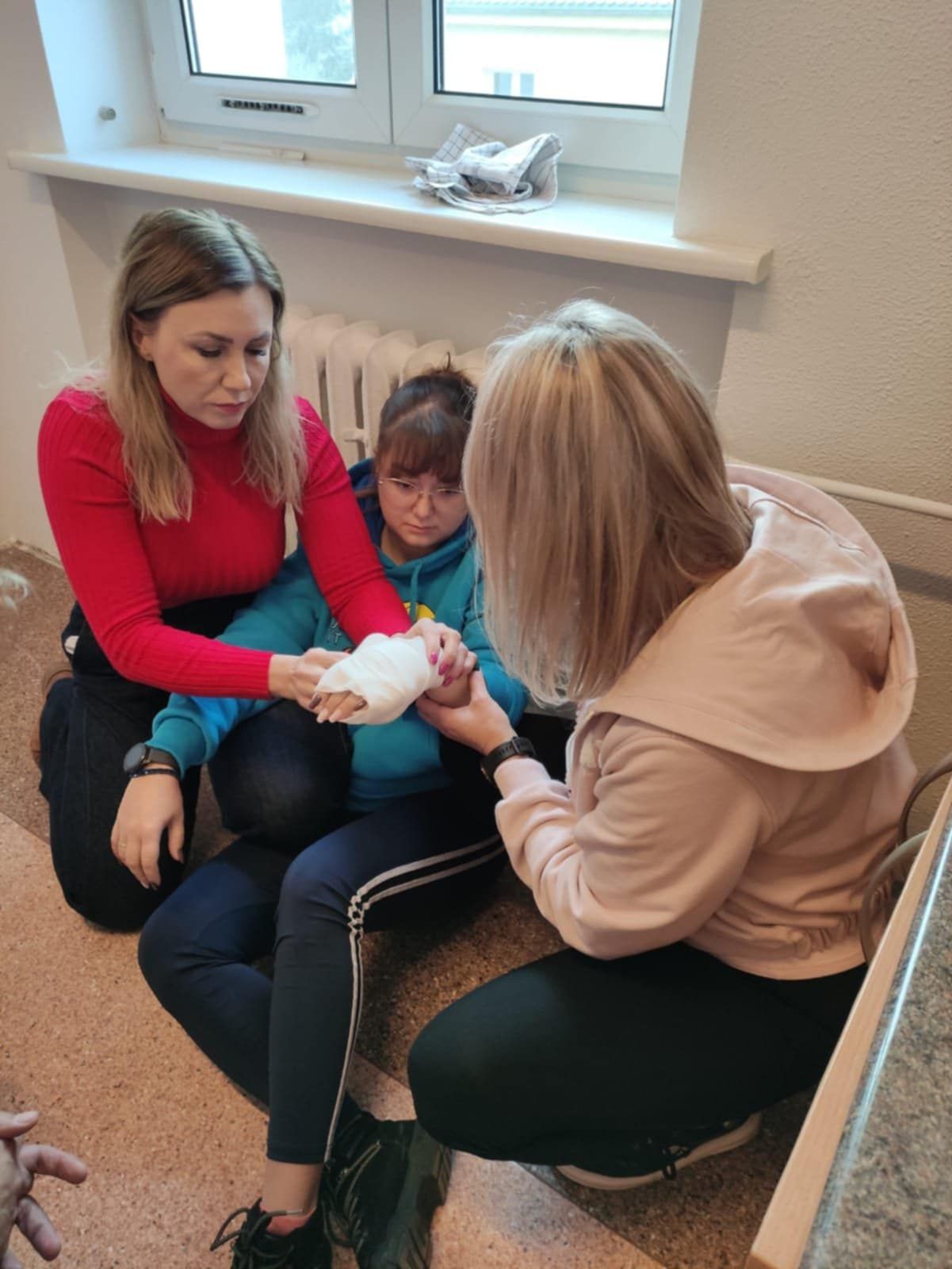Trzy kobiety siedzą na podłodze przy oknie. Dwie z nich opatrują trzecią osobę, która ma zabandażowaną dłoń. Kobieta w czerwonym swetrze podtrzymuje jej ramię, a druga, w jasnej bluzie, poprawia bandaż. Sytuacja wygląda jak ćwiczenia z pierwszej pomocy.