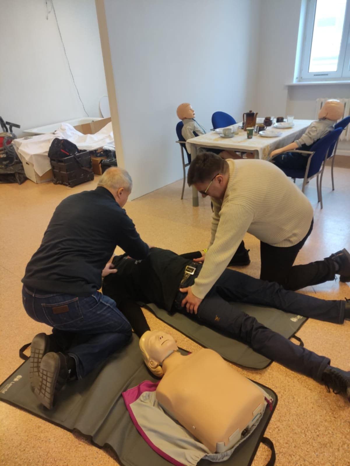 Dwóch mężczyzn udziela pierwszej pomocy osobie leżącej na matach treningowych. W tle stoją manekiny medyczne przy stole, a na podłodze leży także tors manekina do ćwiczeń resuscytacji. Scena przedstawia szkolenie z pierwszej pomocy.