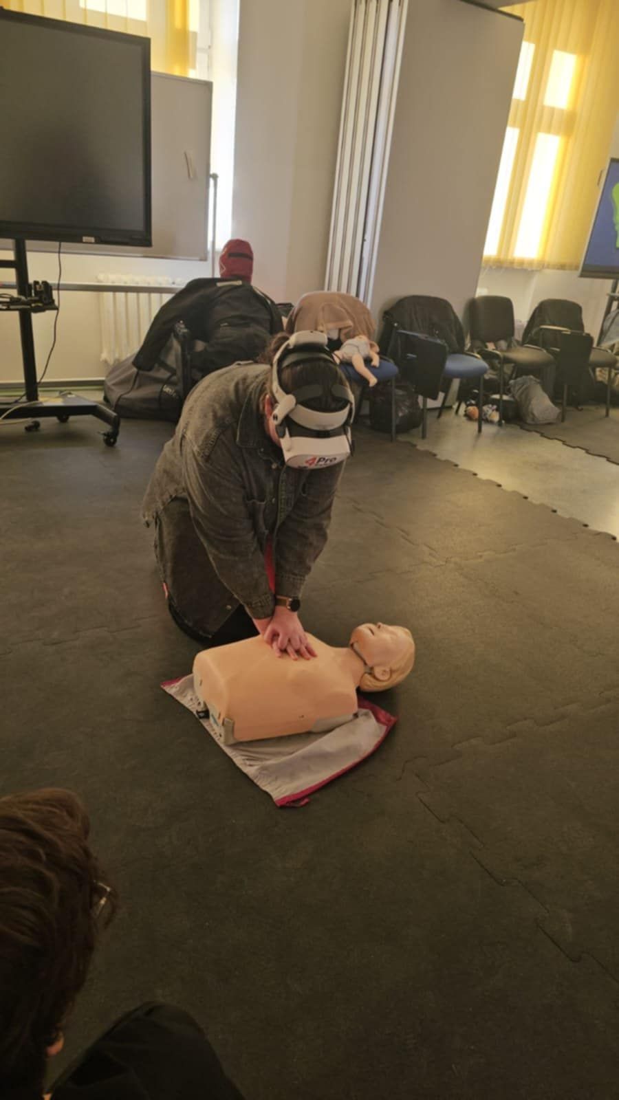 „Studentka w goglach VR przeprowadza uciśnięcia klatki piersiowej na fantomie w sali szkoleniowej wyposażonej w sprzęt medyczny i dydaktyczny.”