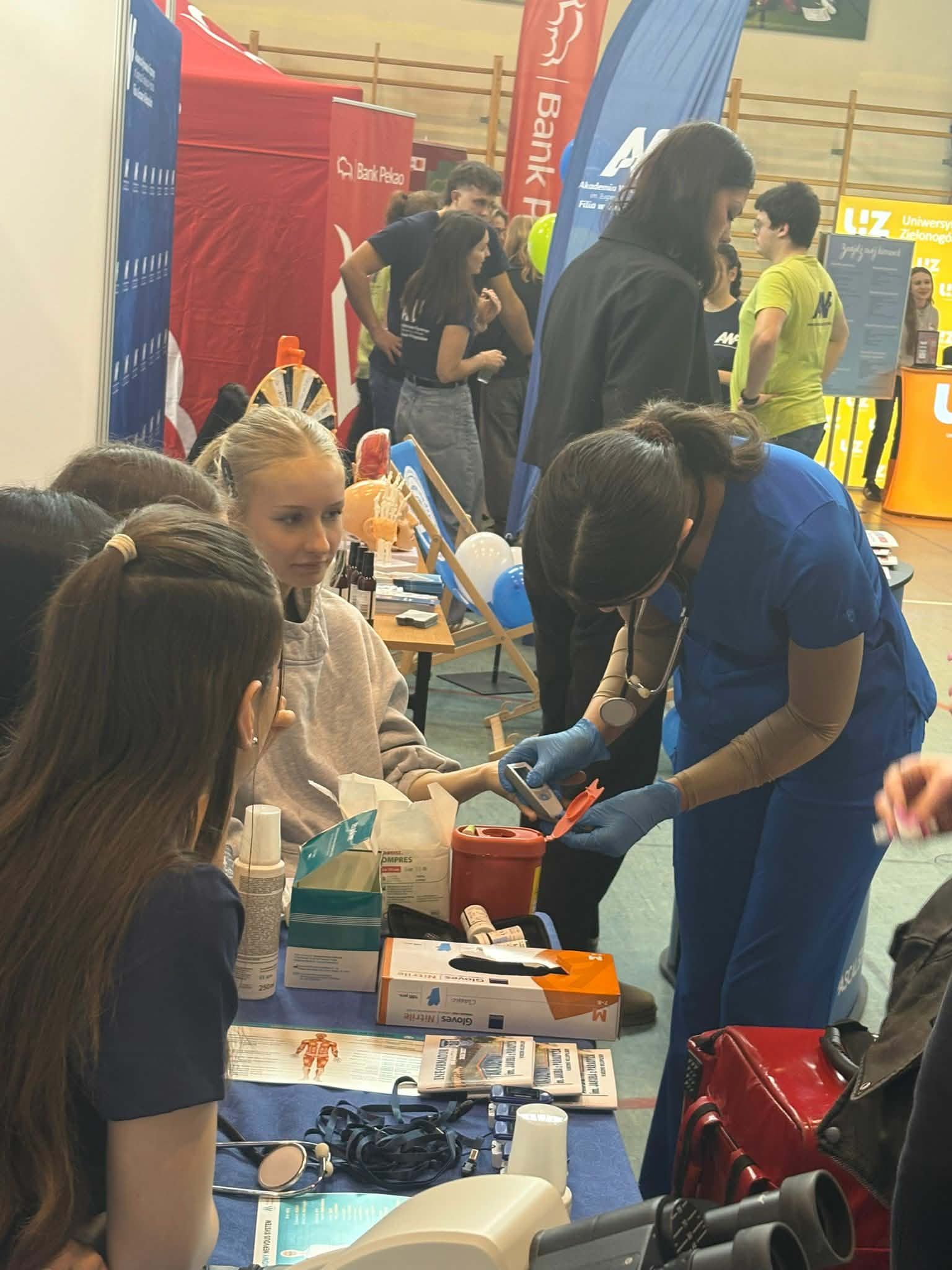 Pokaz medyczny podczas wydarzenia – studentka kierunku pielęgniarstwo wykonuje pomiar poziomu cukru u młodej dziewczyny siedzącej przy stole. Wokół znajdują się materiały medyczne, rękawiczki, pojemniki oraz sprzęt diagnostyczny. Inni uczestnicy obserwują pokaz.