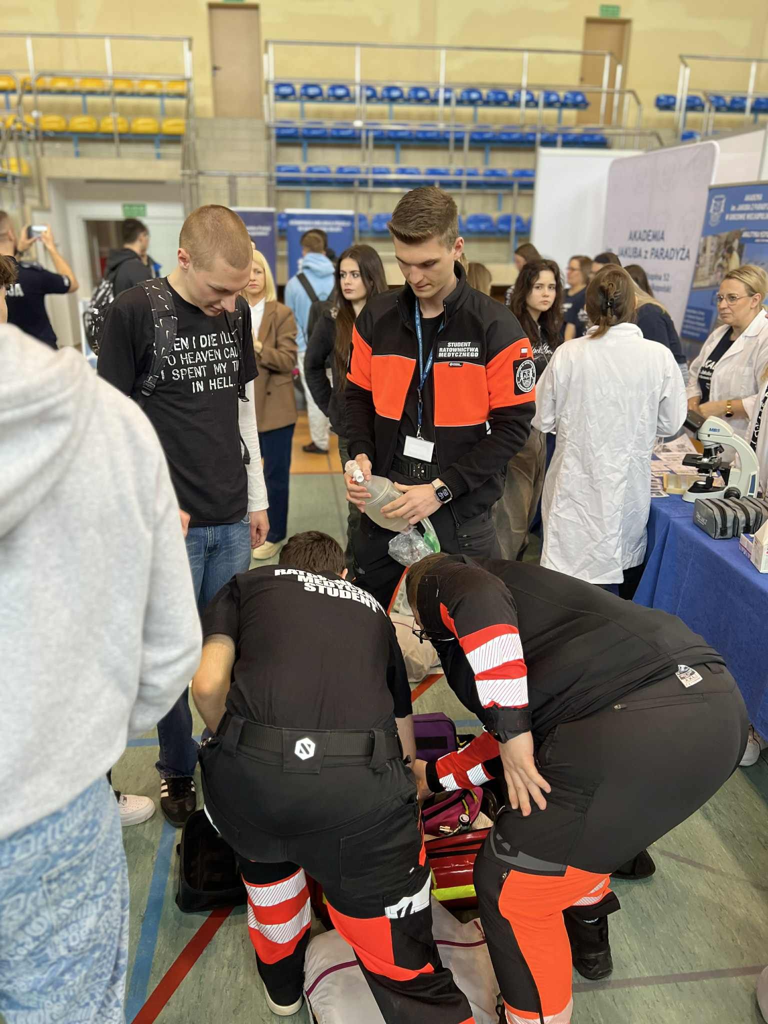 Studenci kierunku ratownictwo medyczne w czarno-pomarańczowych uniformach udzielają pomocy na fantomie leżącym na podłodze w hali sportowej. Jeden z nich przygotowuje sprzęt do udrażniania dróg oddechowych, a inni pochylają się nad fantomem. Wokół stoją obserwatorzy, przyglądając się pokazowi pierwszej pomocy.