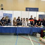 Grupa studentów i dydaktyków stoi przy stoisku Akademii. Na stołach znajdują się materiały medyczne, probówki i mikroskop. Część osób ma na sobie koszulki uczelni, a w tle widoczne są banery promujące kierunki związane ze zdrowiem. Na podłodze leży fantom do nauki pierwszej pomocy oraz torba ratownicza.