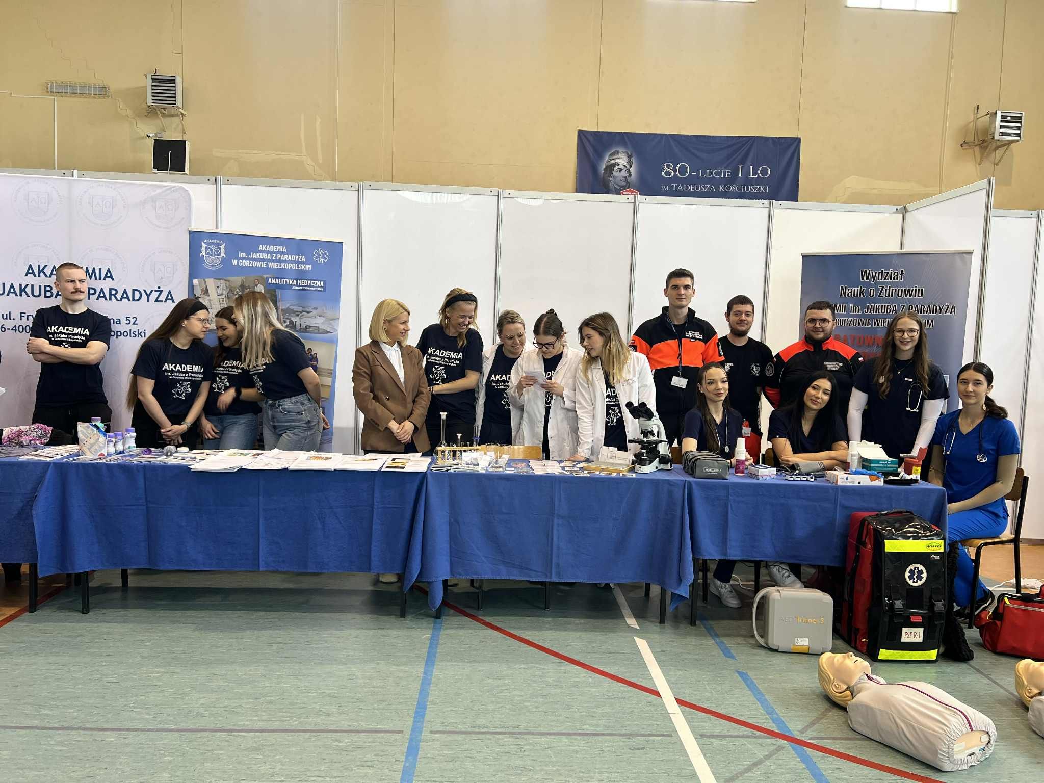 Grupa studentów i dydaktyków stoi przy stoisku Akademii. Na stołach znajdują się materiały medyczne, probówki i mikroskop. Część osób ma na sobie koszulki uczelni, a w tle widoczne są banery promujące kierunki związane ze zdrowiem. Na podłodze leży fantom do nauki pierwszej pomocy oraz torba ratownicza.