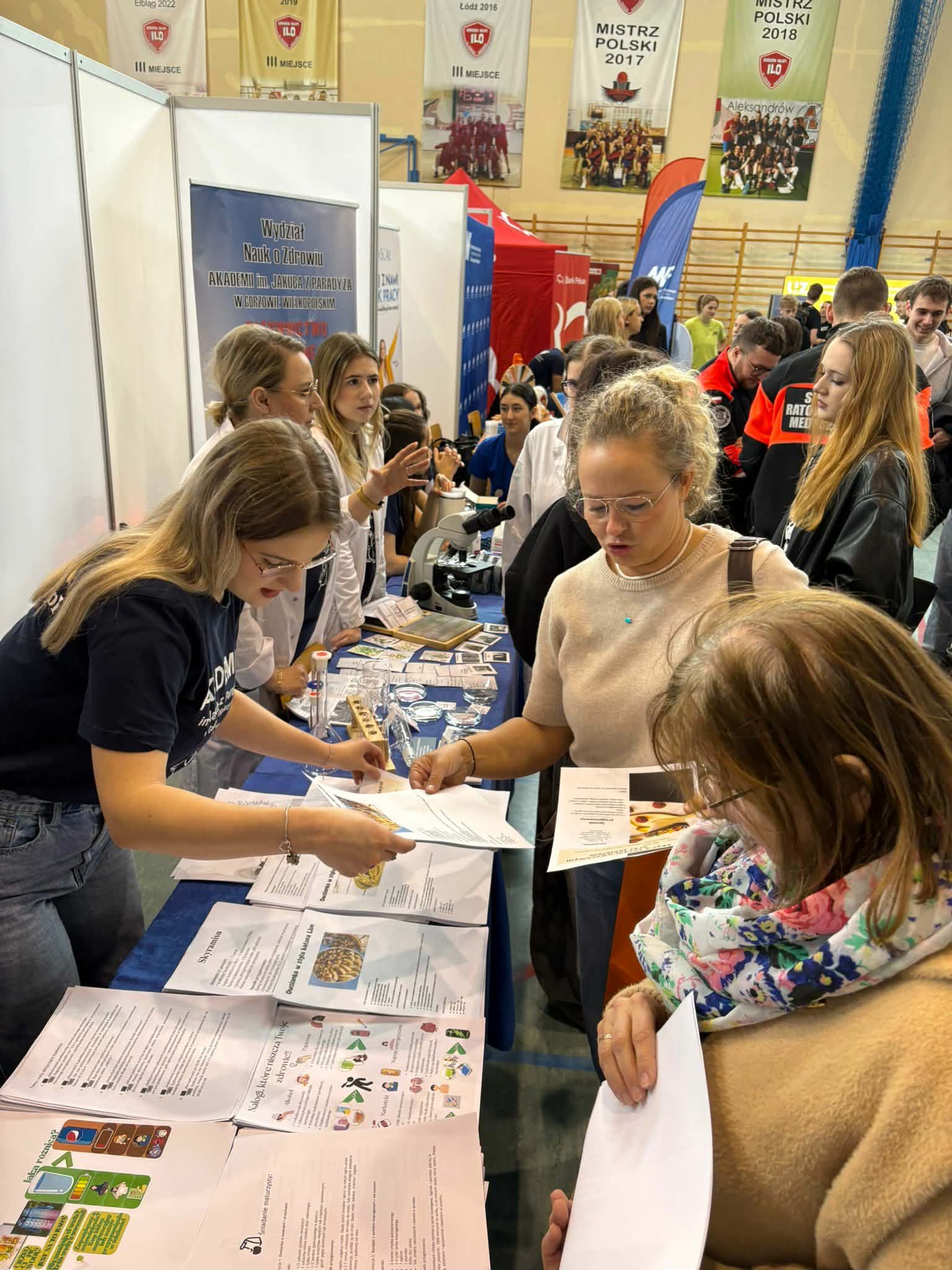 Stoisko informacyjne na targach edukacyjnych w hali sportowej. Studentki rozmawiają z odwiedzającymi i rozdają ulotki oraz materiały informacyjne. Na stole leżą broszury, plansze edukacyjne i mikroskop. W tle widać inne stoiska oraz bannery uczelni.
