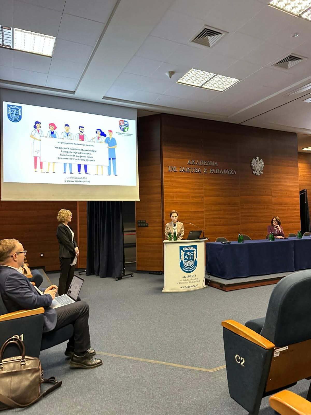 Pani Rektor Akademii im. Jakuba z Paradyża prof. dr hab. Elżbieta Skorupska-Raczyńska przemawia przy mównicy podczas konferencji w sali wykładowej, a na ekranie wyświetlona jest prezentacja dotycząca ochrony zdrowia.