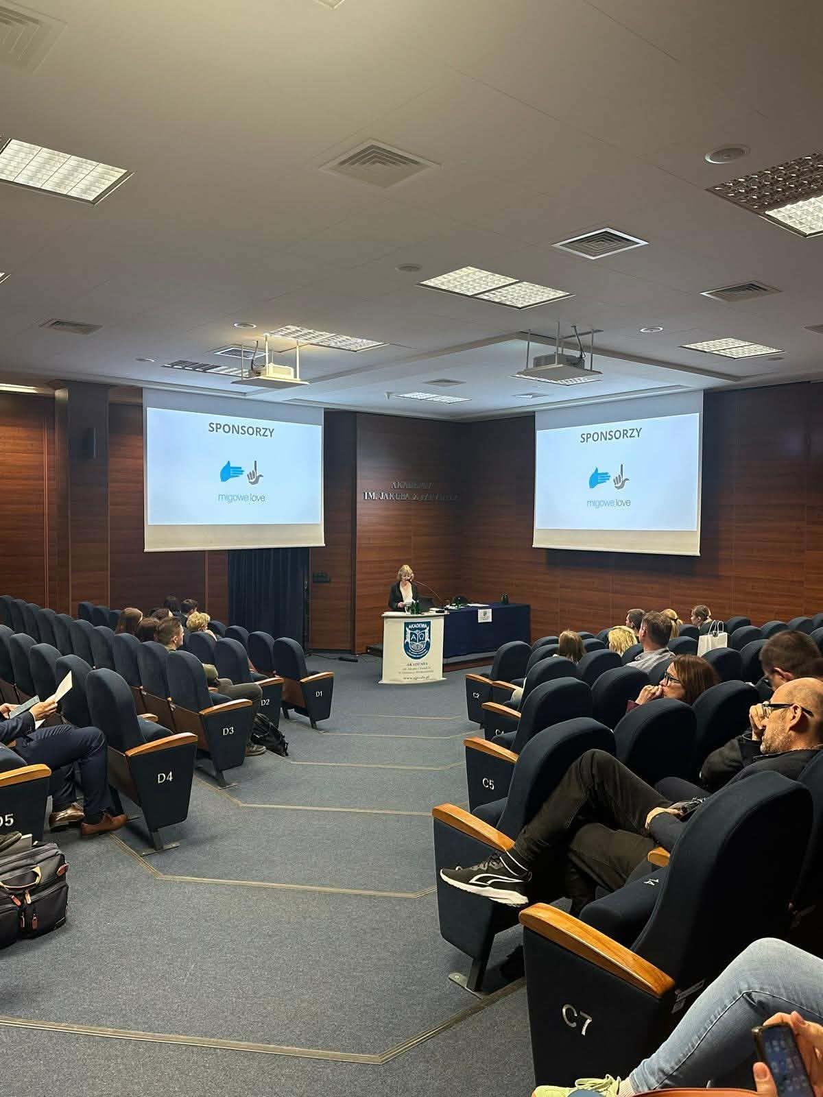 Sala konferencyjna z rzędami granatowych foteli i uczestnikami siedzącymi w trakcie prezentacji. Na środku widoczna pani dziekan Wydziału Nauk o Zdrowiu prof. AJP dr Dorota Skrocka stojąca przy mównicy, a po obu stronach wyświetlane slajdy z napisem „Sponsorzy”.