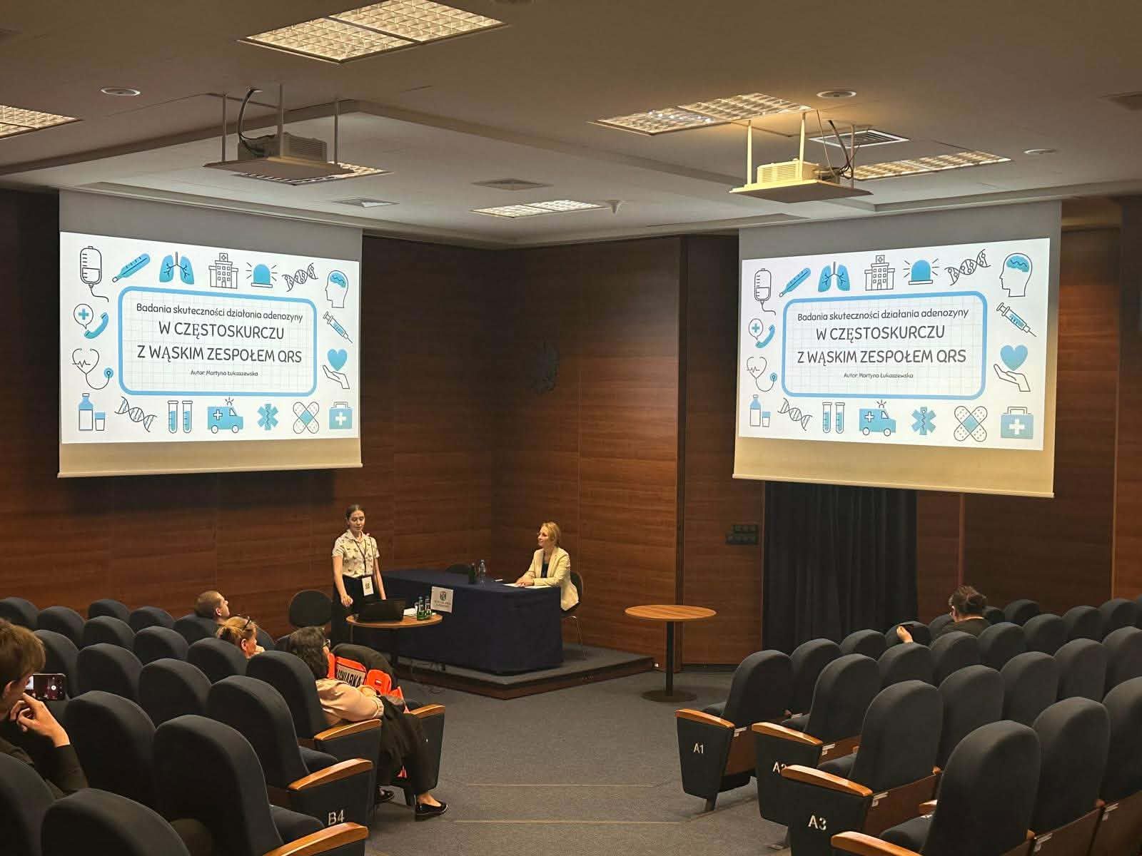 Wykład medyczny w nowoczesnej sali – na dwóch ekranach wyświetlany slajd dotyczący badań nad działaniem adenozyny w częstoskurczu. Jedna osoba prezentuje, druga siedzi przy stole, a publiczność obserwuje wystąpienie.