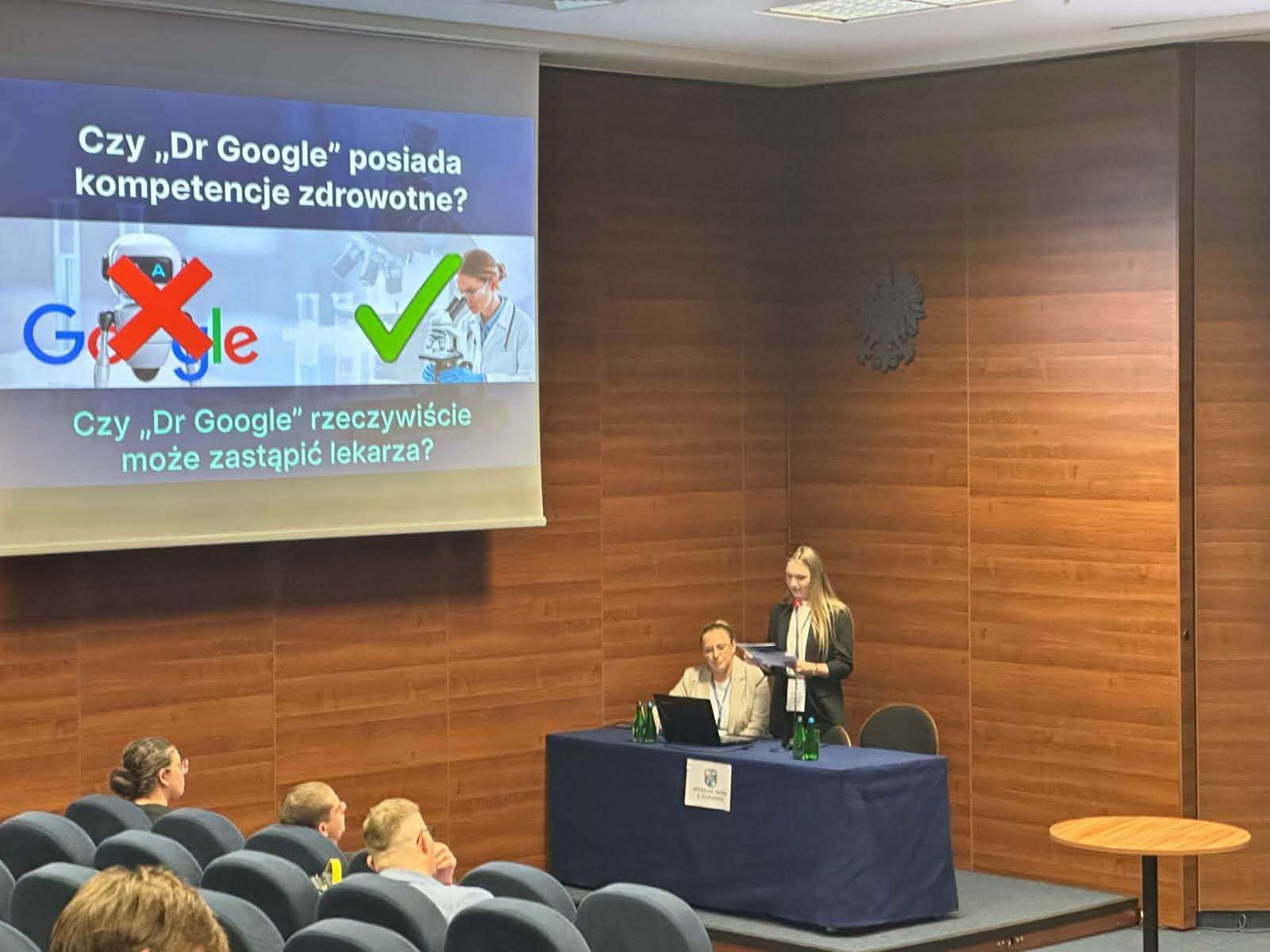 Prelegentka stoi obok siedzącej kobiety przy stole i prowadzi prezentację. Na ekranie slajd o tym, czy „Dr Google” może zastąpić lekarza.
