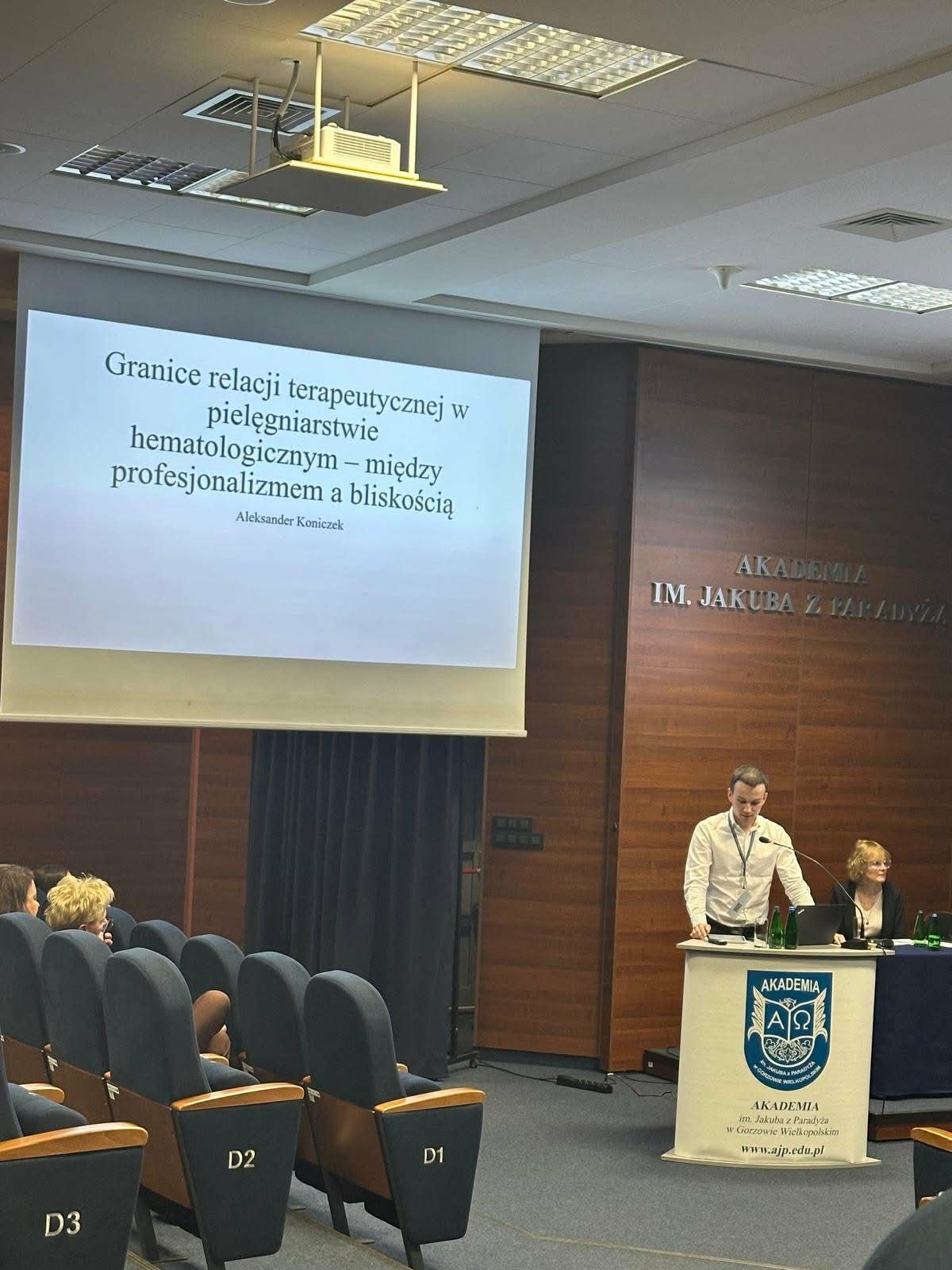 Mężczyzna stoi przy mównicy i wygłasza prezentację w sali wykładowej. Na ekranie widoczny jest slajd o granicach relacji terapeutycznej w pielęgniarstwie hematologicznym. Na sali siedzi publiczność.