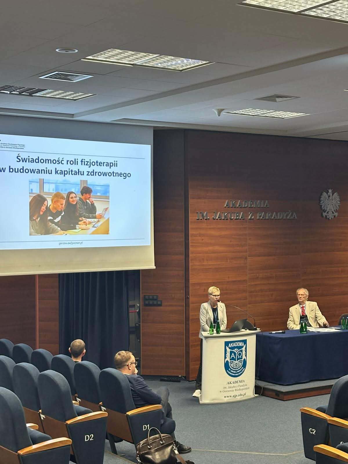 Sala wykładowa z prelegentką przy mównicy. Na ekranie prezentacja o roli fizjoterapii w budowaniu kapitału zdrowotnego.