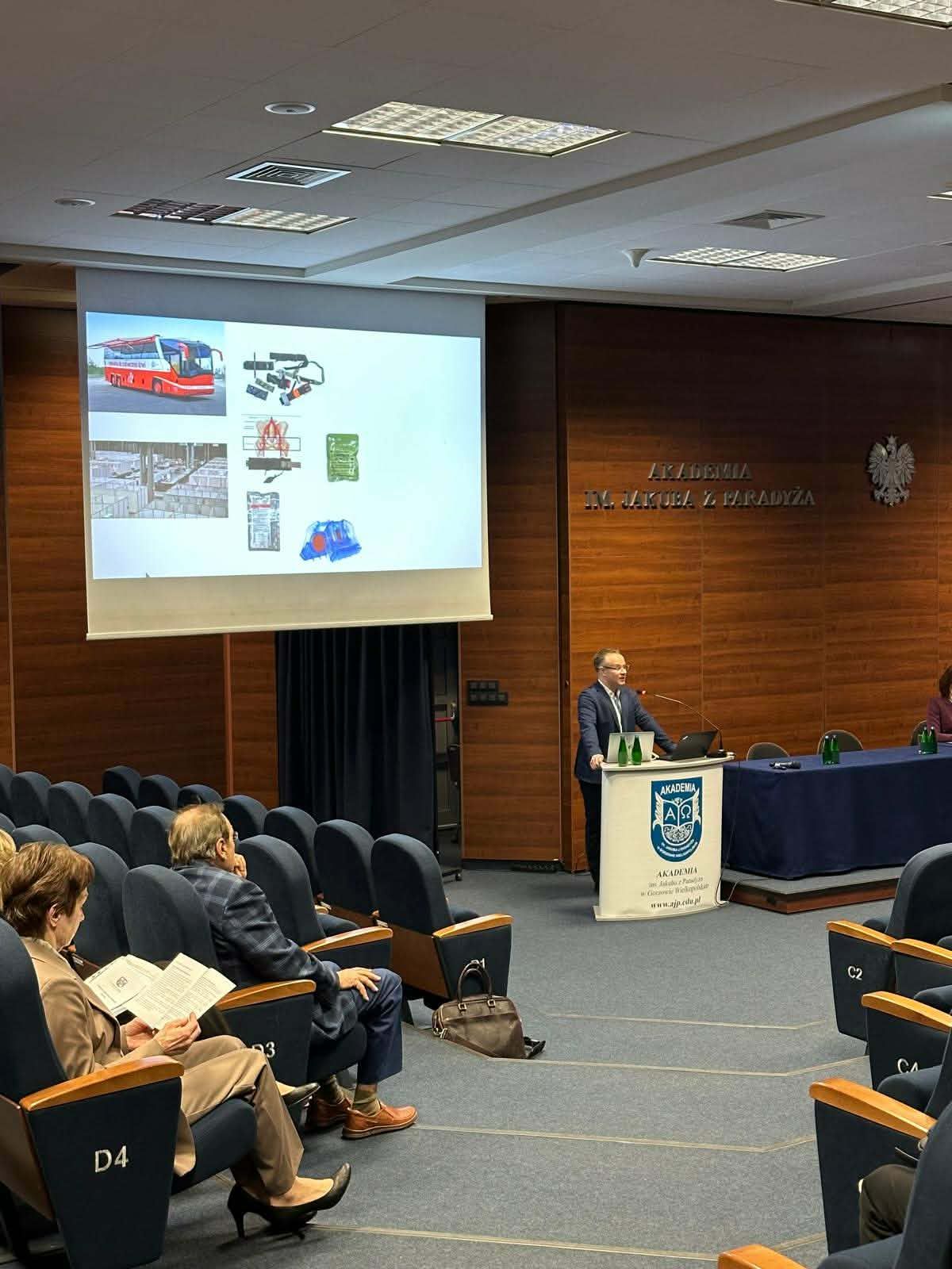 Mężczyzna prowadzi wykład w sali konferencyjnej. Na ekranie widoczna prezentacja z ilustracjami pojazdów i urządzeń, a uczestnicy siedzą w rzędach i słuchają.