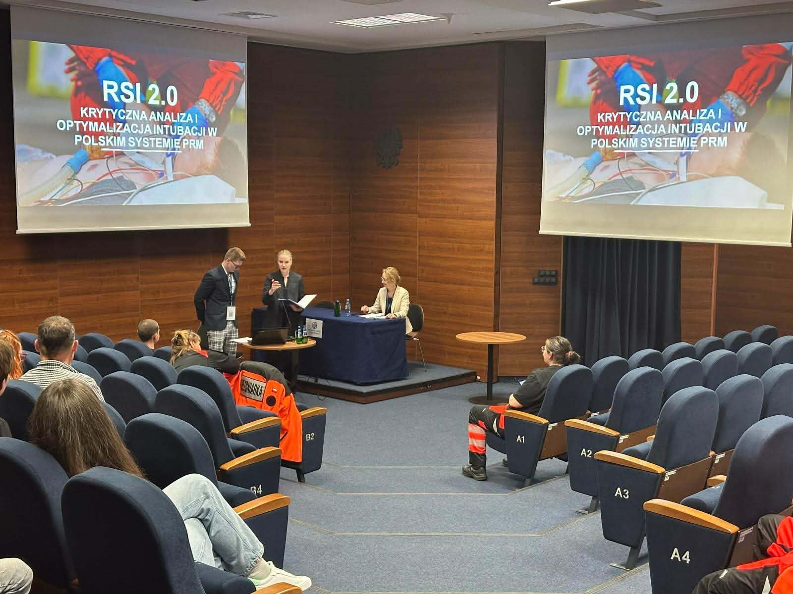 Sala konferencyjna z uczestnikami siedzącymi w rzędach foteli. Z przodu dwie osoby prowadzą prezentację, a na dwóch ekranach wyświetlony jest slajd „RSI 2.0” dotyczący analizy i optymalizacji intubacji w polskim systemie ratownictwa medycznego.